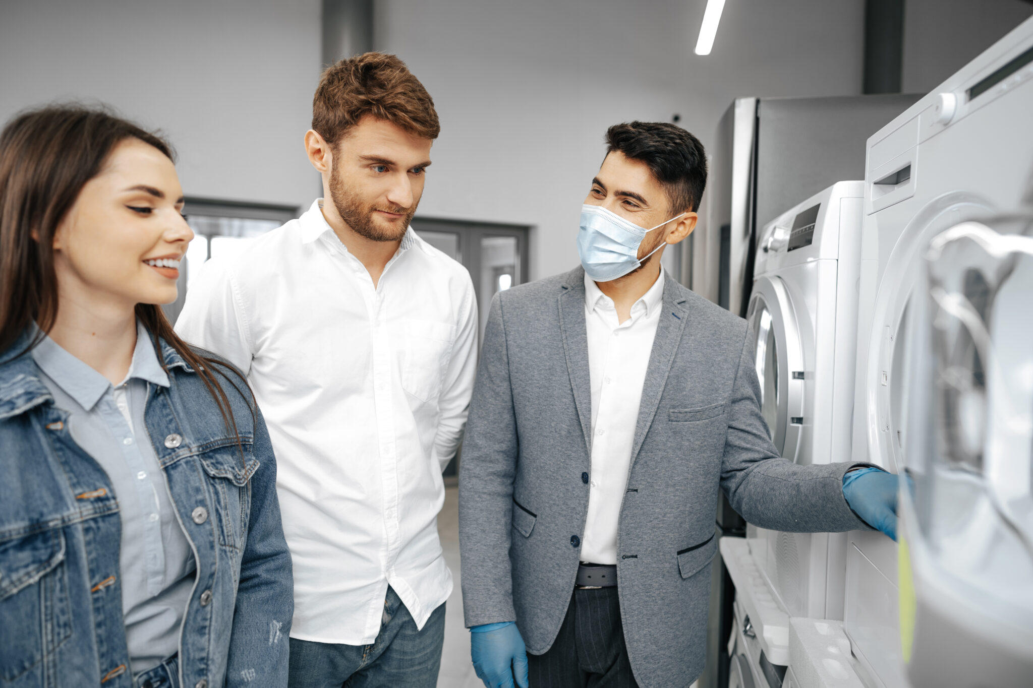 Salesman in hypermarket wearing medical mask demonstrates his clients a new washing machine, close up